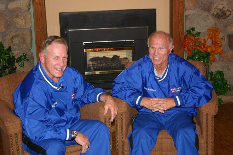Two men in blue Mile High Karate jackets seated by a fireplace; the jacket on the right is embroidered Bill Wallace