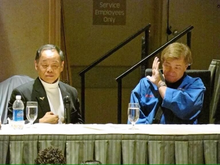 Grand Master Jhoon Rhee seated at a head table beside a Black Belt in a blue uniform at the Mile High Karate Black Belt Retreat