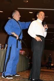 Grand Master Jhoon Rhee speaking with a microphone beside a Black Belt in a blue uniform at the Mile High Karate Black Belt Retreat