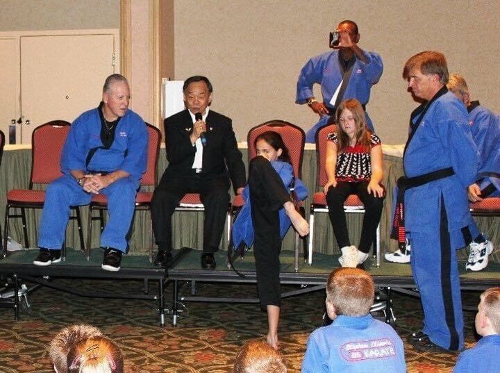 Grand Master Jhoon Rhee on stage with a microphone while a young student demonstrates a kick at the Mile High Karate Black Belt Retreat